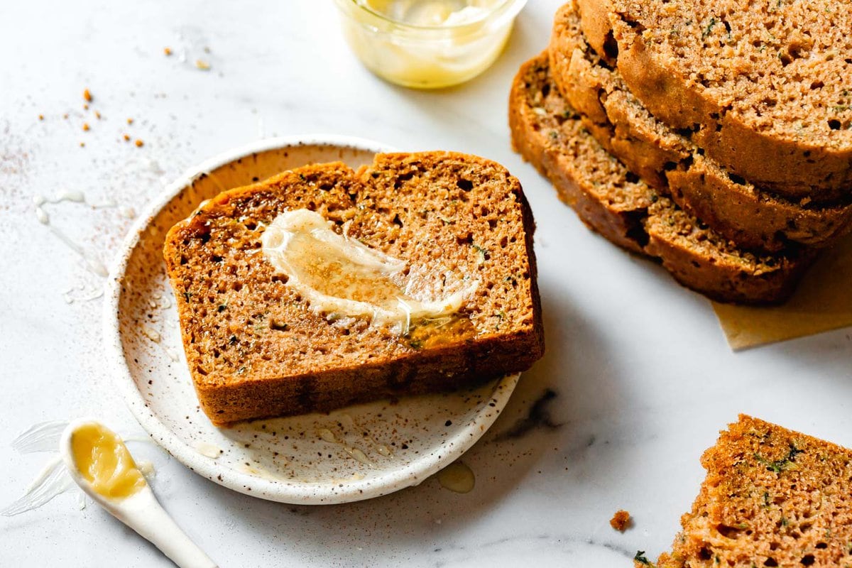 Zucchini Bread — Moist Cinnamon Spiced Quick Bread You'll Make All Summer 28 Close-up of a single slice of zucchini bread on a white plate, showing the moist green-flecked crumb, with a smear of almond butter and a cup of tea in soft focus background