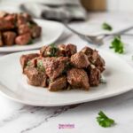 Air fryer steak bites piled on a white plate, garnished with fresh parsley and garlic butter, shot on a dark wood surface