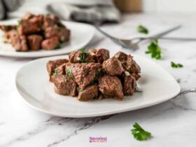 Air fryer steak bites piled on a white plate, garnished with fresh parsley and garlic butter, shot on a dark wood surface