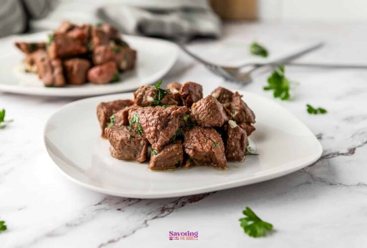 Air fryer steak bites piled on a white plate, garnished with fresh parsley and garlic butter, shot on a dark wood surface