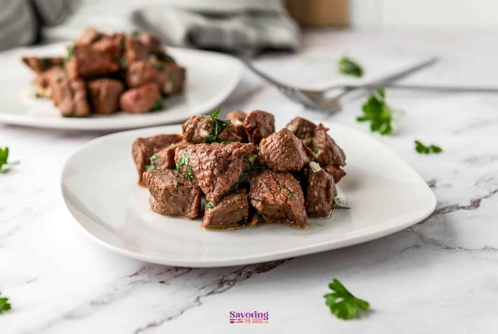 Air fryer steak bites piled on a white plate, garnished with fresh parsley and garlic butter, shot on a dark wood surface