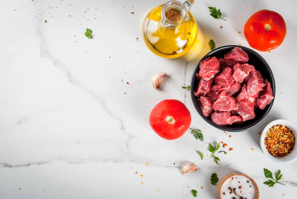 Raw sirloin steak cubes in a glass bowl being tossed with garlic, smoked paprika, and Worcestershire sauce, overhead shot on a marble counter