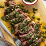 Argentinian chimichurri steak sliced on a wooden board with bright green chimichurri sauce drizzled over the top, fresh parsley garnish, and a rustic linen napkin beside it