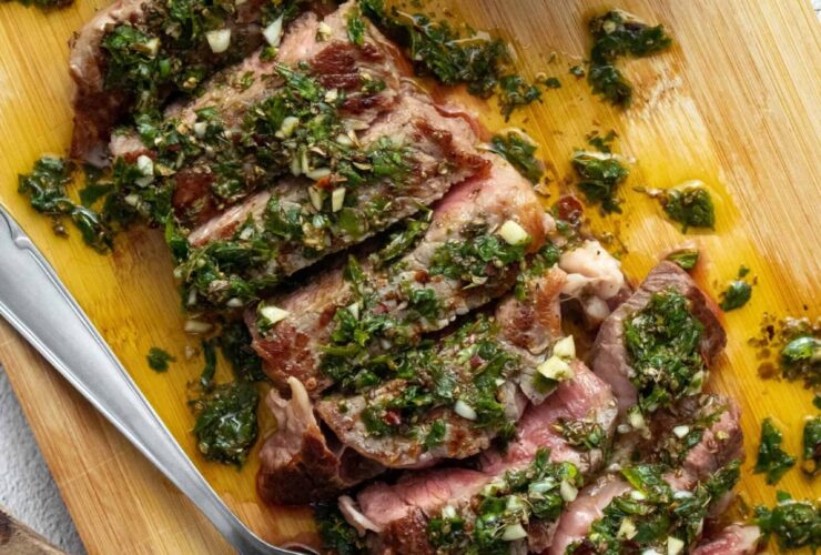 Argentinian chimichurri steak sliced on a wooden board with bright green chimichurri sauce drizzled over the top, fresh parsley garnish, and a rustic linen napkin beside it