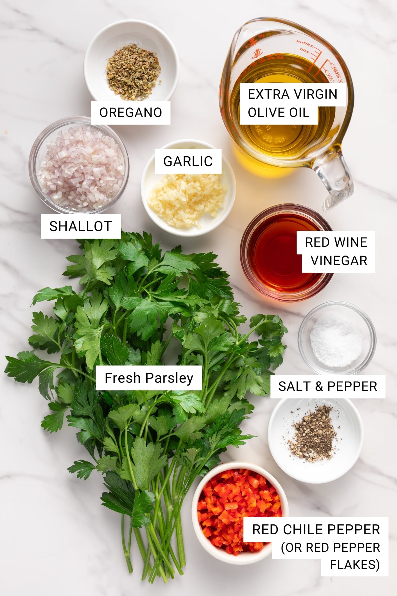 Overhead flat lay of chimichurri ingredients including fresh parsley, garlic cloves, red wine vinegar bottle, olive oil, dried red pepper flakes, and fresh oregano on a white marble surface