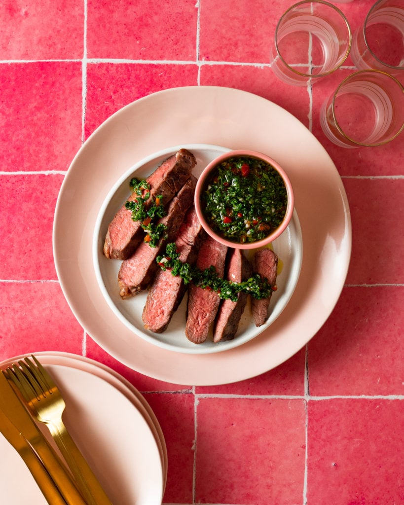 Close-up of sliced Argentinian chimichurri steak fanned out on a white plate, glossy chimichurri pooling over the meat, showing the juicy pink interior and vibrant green herb sauce