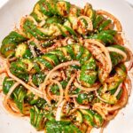 Overhead shot of Asian spiralized cucumber noodle salad in a wide bowl, garnished with sesame seeds, sliced scallions, shredded carrots, and a drizzle of sesame-ginger dressing on a light linen surface