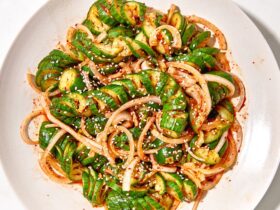 Overhead shot of Asian spiralized cucumber noodle salad in a wide bowl, garnished with sesame seeds, sliced scallions, shredded carrots, and a drizzle of sesame-ginger dressing on a light linen surface