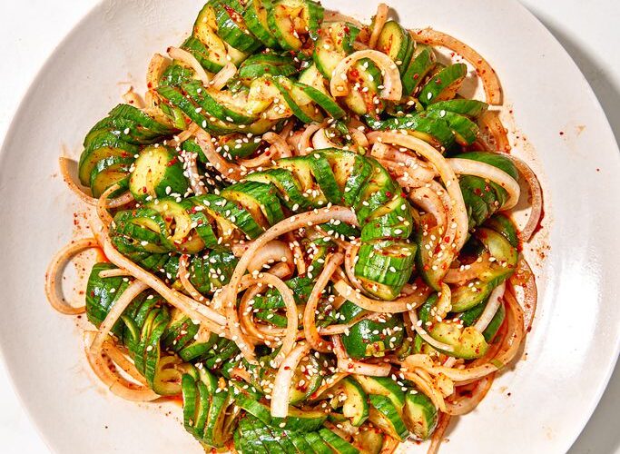 Overhead shot of Asian spiralized cucumber noodle salad in a wide bowl, garnished with sesame seeds, sliced scallions, shredded carrots, and a drizzle of sesame-ginger dressing on a light linen surface