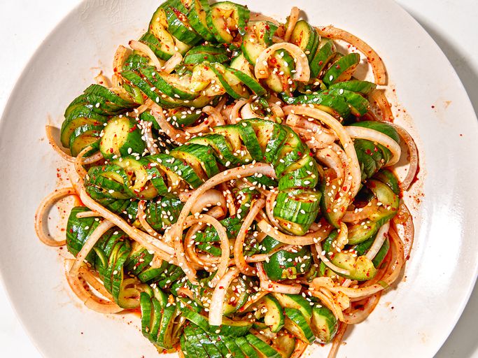 Overhead shot of Asian spiralized cucumber noodle salad in a wide bowl, garnished with sesame seeds, sliced scallions, shredded carrots, and a drizzle of sesame-ginger dressing on a light linen surface