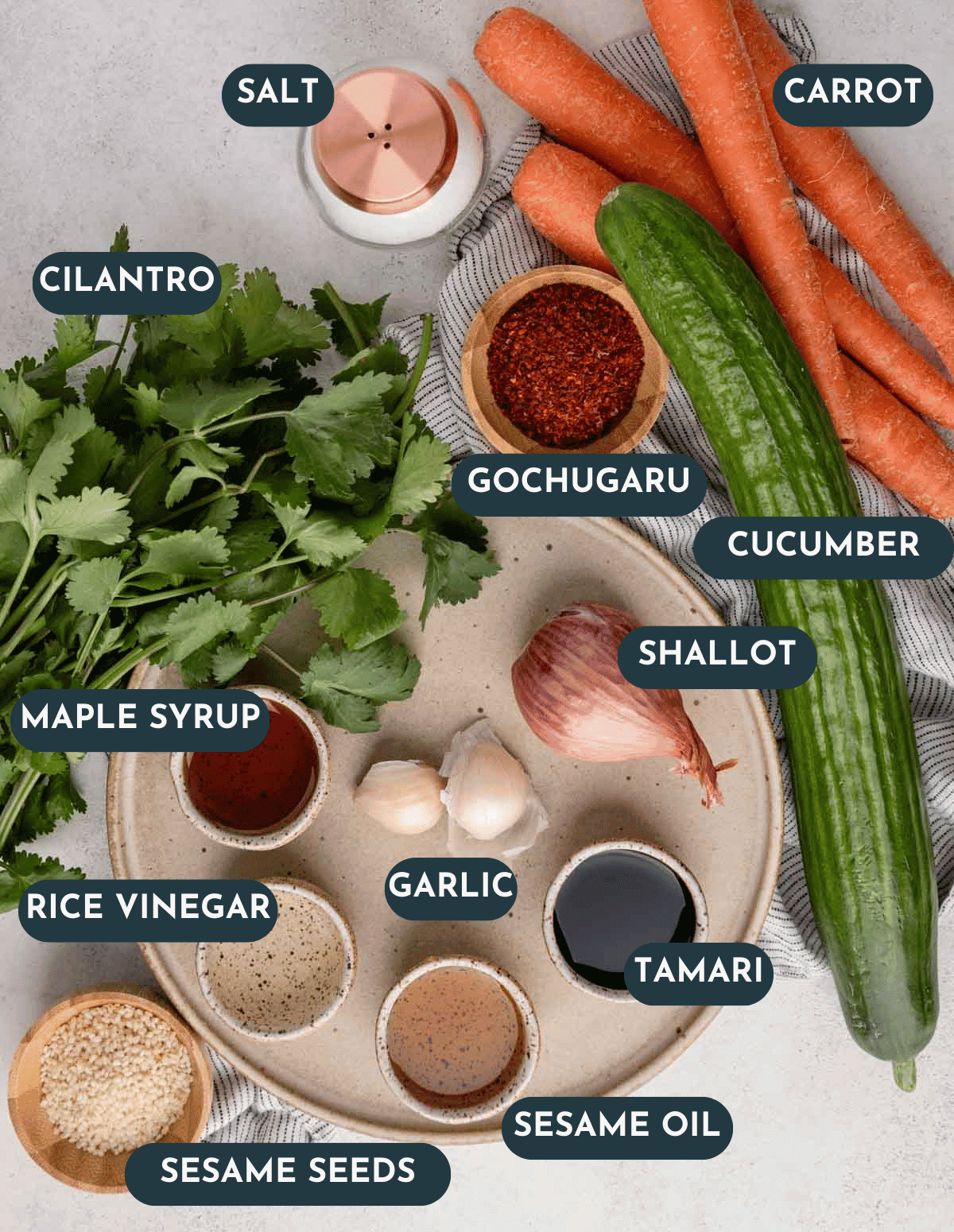 Flat-lay of ingredients for Asian cucumber noodle salad including English cucumbers, fresh ginger root, toasted sesame oil, tamari, rice vinegar, scallions, carrots, sesame seeds, and a spiralizer on a white marble surface