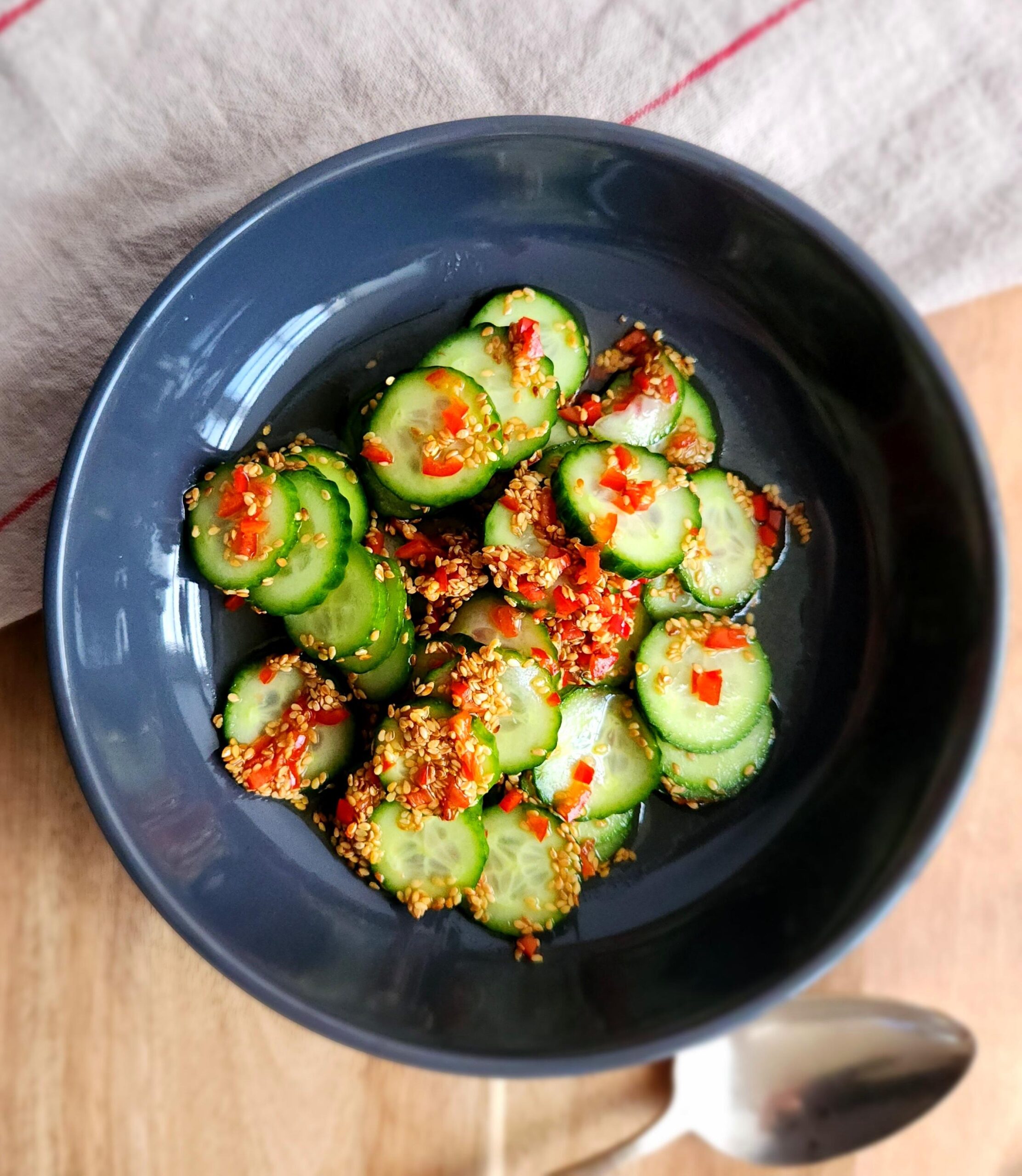 Close-up shot of spiralized cucumber noodle salad in a black bowl, showing the glistening sesame-ginger dressing coating the cucumber noodles, with sesame seeds, red chili flakes, and fresh cilantro garnish in soft natural light