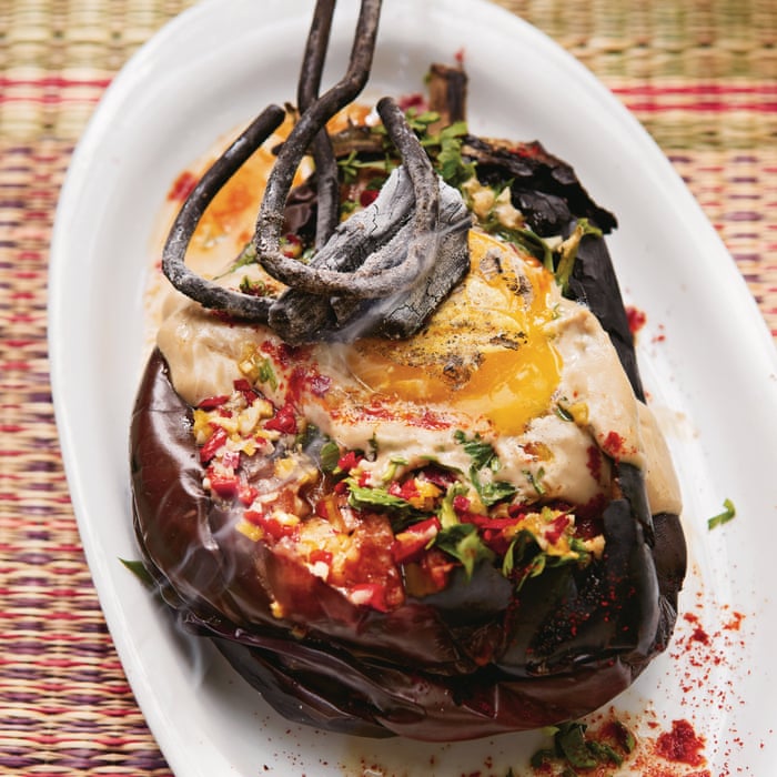 Two large globe eggplants charring directly over a gas burner flame alongside small bowls of tahini, lemon, garlic, and olive oil arranged on a marble surface