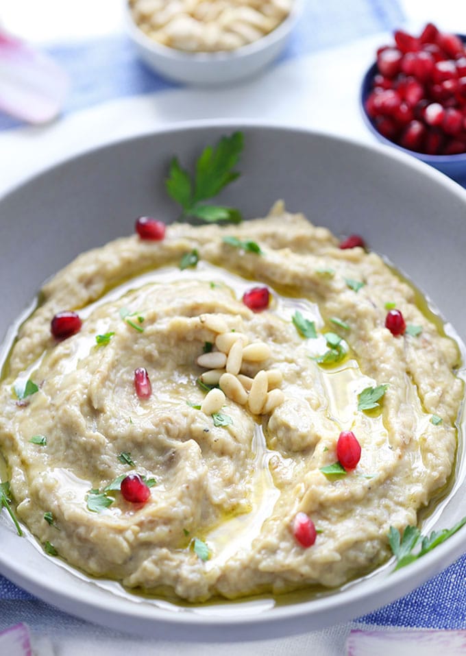 Close-up overhead shot of Baba Ganoush in a white ceramic bowl with a swirl of olive oil, pomegranate arils scattered across the surface, and warm toasted pita triangles leaning against the bowl