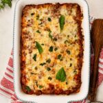 golden bubbly baked ziti in a white ceramic baking dish, fresh basil on top, rustic wooden table