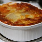 golden bubbly baked ziti in a white ceramic baking dish, fresh basil on top, rustic wooden table