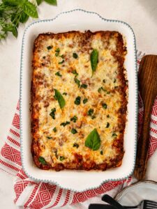 golden bubbly baked ziti in a white ceramic baking dish, fresh basil on top, rustic wooden table