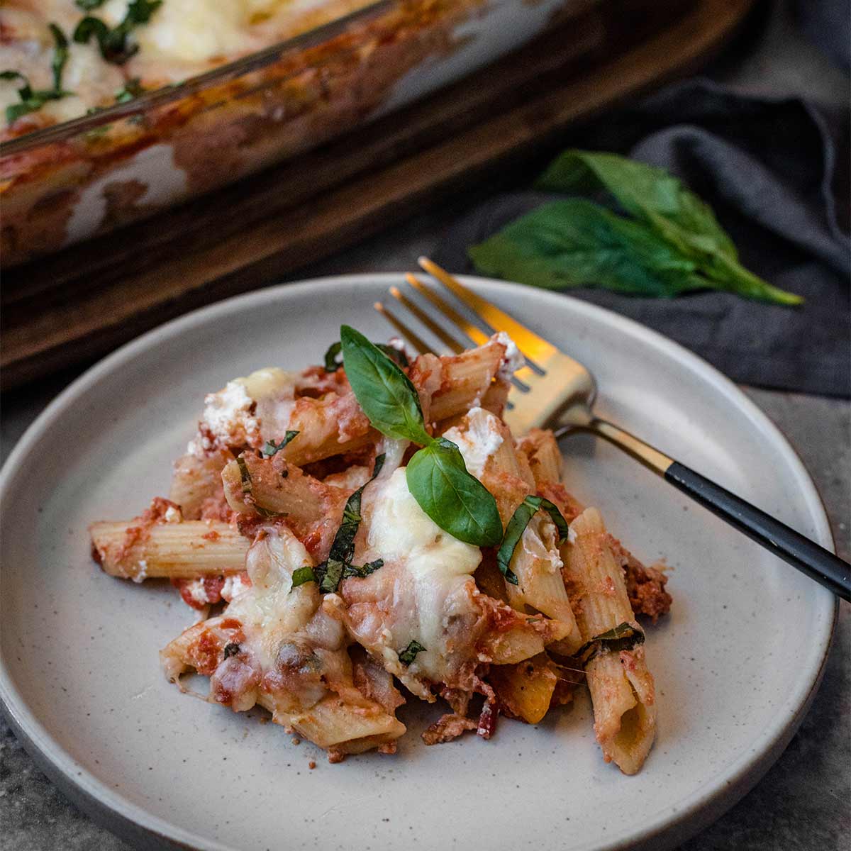 overhead flat lay of baked ziti ingredients including raw pasta, ricotta in a bowl, shredded mozzarella, crushed tomatoes, and fresh herbs on a marble surface