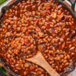 A large cast iron skillet filled with glossy, smoky BBQ baked beans garnished with crispy bacon crumbles and a sprig of fresh thyme, photographed on a rustic wooden outdoor table surrounded by summer cookout props