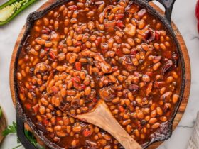 A large cast iron skillet filled with glossy, smoky BBQ baked beans garnished with crispy bacon crumbles and a sprig of fresh thyme, photographed on a rustic wooden outdoor table surrounded by summer cookout props