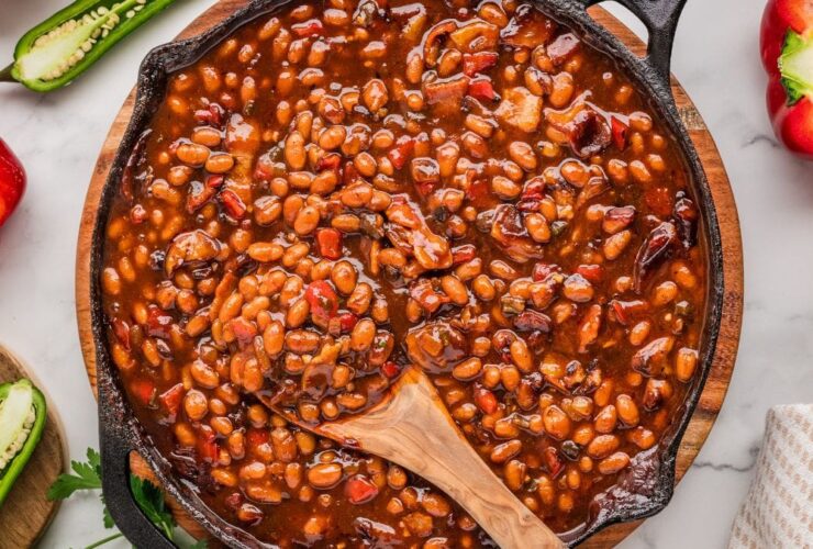 A large cast iron skillet filled with glossy, smoky BBQ baked beans garnished with crispy bacon crumbles and a sprig of fresh thyme, photographed on a rustic wooden outdoor table surrounded by summer cookout props