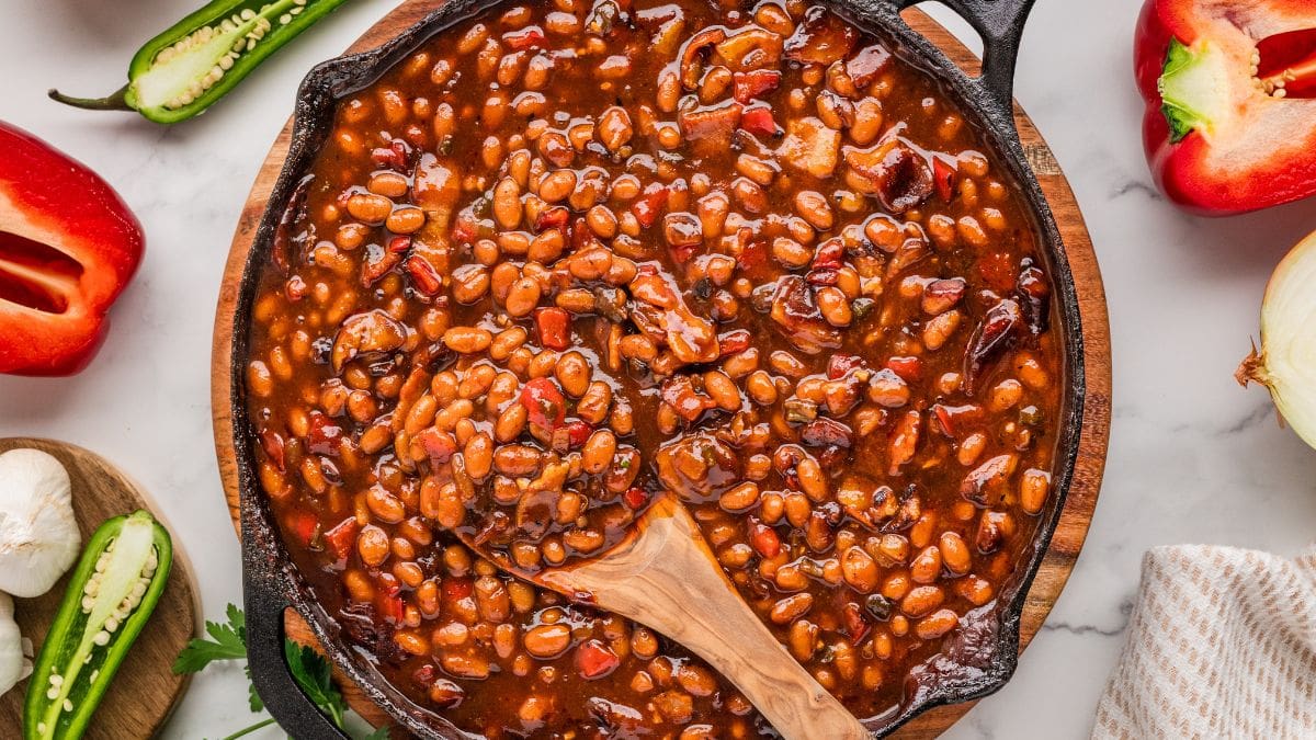 BBQ Baked Beans — Smoky Sweet 4th of July Side Dish 26 A large cast iron skillet filled with glossy, smoky BBQ baked beans garnished with crispy bacon crumbles and a sprig of fresh thyme, photographed on a rustic wooden outdoor table surrounded by summer cookout props
