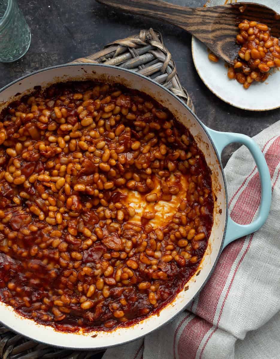 BBQ Baked Beans — Smoky Sweet 4th of July Side Dish 27 A flat lay overhead shot of all BBQ baked beans ingredients laid out on a white marble surface — cans of navy beans, thick-cut bacon strips, a bowl of brown sugar, smoked paprika, apple cider vinegar, tomato paste, onion, and garlic cloves