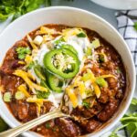A deep bowl of thick beef chili topped with shredded cheddar, sour cream, sliced jalapeños, and green onions, photographed from above on a rustic wooden table with a side of cornbread