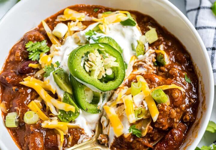 A deep bowl of thick beef chili topped with shredded cheddar, sour cream, sliced jalapeños, and green onions, photographed from above on a rustic wooden table with a side of cornbread