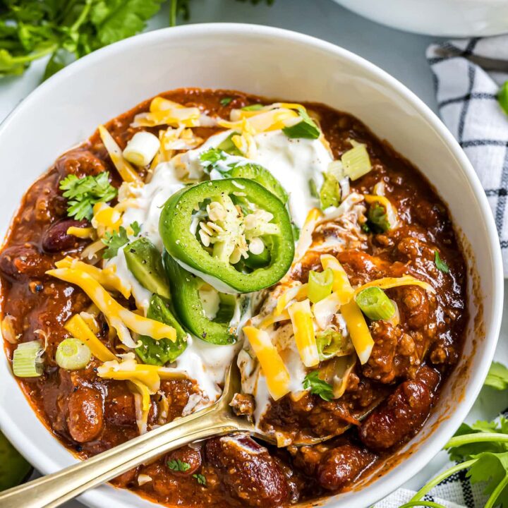 A deep bowl of thick beef chili topped with shredded cheddar, sour cream, sliced jalapeños, and green onions, photographed from above on a rustic wooden table with a side of cornbread