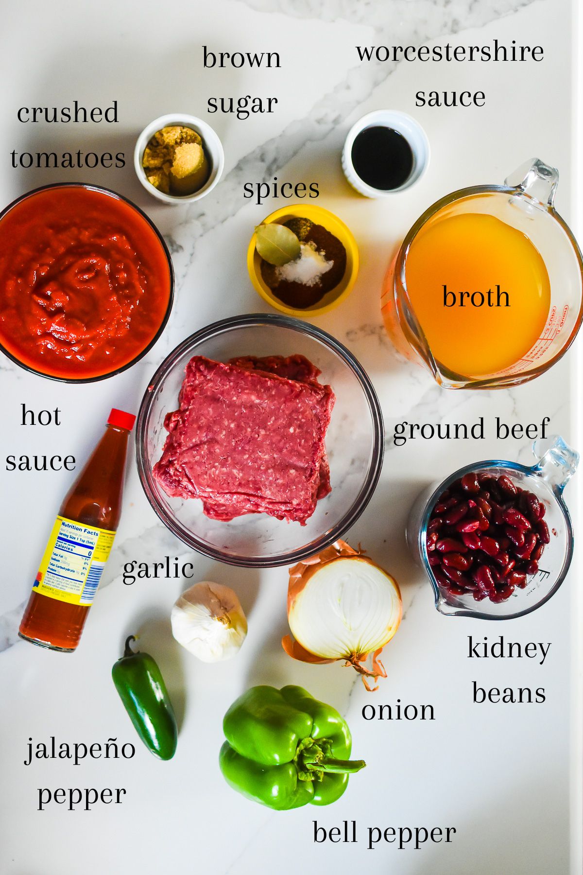 An overhead flat-lay of all beef chili ingredients prepped and laid out — browned ground beef in a skillet, drained kidney beans, canned fire-roasted tomatoes, diced onions, minced garlic, and small bowls of chili spices on a marble surface