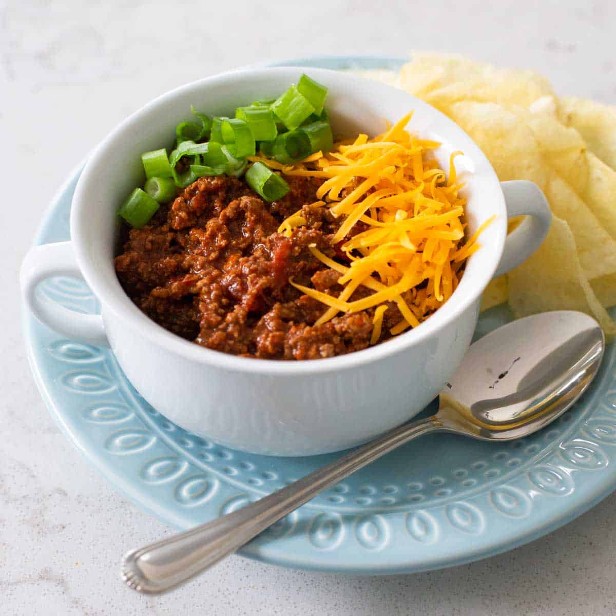 A close-up side shot of a white ceramic bowl of beef chili topped with a generous mound of shredded cheddar cheese melting into the hot chili, with a sprig of cilantro and a lime wedge on the side