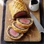 A perfectly sliced British Beef Wellington on a white marble serving board, revealing a rosy pink beef center surrounded by dark mushroom duxelles and golden flaky puff pastry, garnished with fresh thyme
