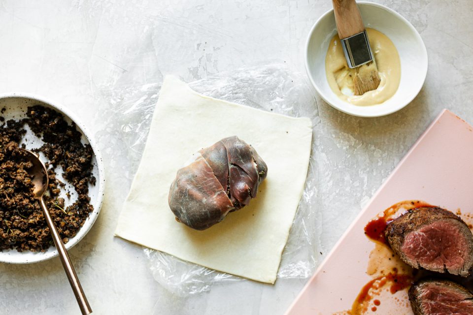 Overhead flat-lay of beef Wellington preparation steps showing seared tenderloin, mushroom duxelles in a pan, prosciutto laid out on plastic wrap, and rolled puff pastry on a baking sheet dusted with flour