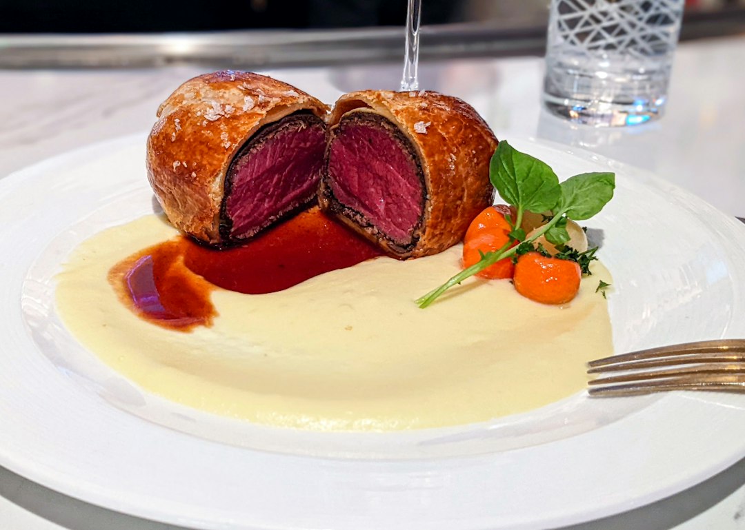 A close-up cross-section slice of British Beef Wellington on a white dinner plate showing the rosy medium-rare beef center, dark green mushroom duxelles layer, thin prosciutto, and shatteringly crisp golden puff pastry exterior, with a side of red wine reduction drizzled on the plate