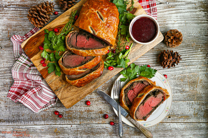A close-up cross-section slice of British Beef Wellington on a white dinner plate showing the rosy medium-rare beef center, dark green mushroom duxelles layer, thin prosciutto, and shatteringly crisp golden puff pastry exterior, with a side of red wine reduction drizzled on the plate
