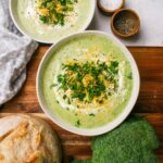 A rustic bowl of broccoli cheddar soup garnished with shredded cheddar and broccoli florets, photographed on a wooden table with warm natural light