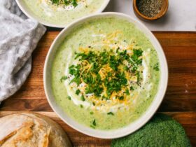 A rustic bowl of broccoli cheddar soup garnished with shredded cheddar and broccoli florets, photographed on a wooden table with warm natural light