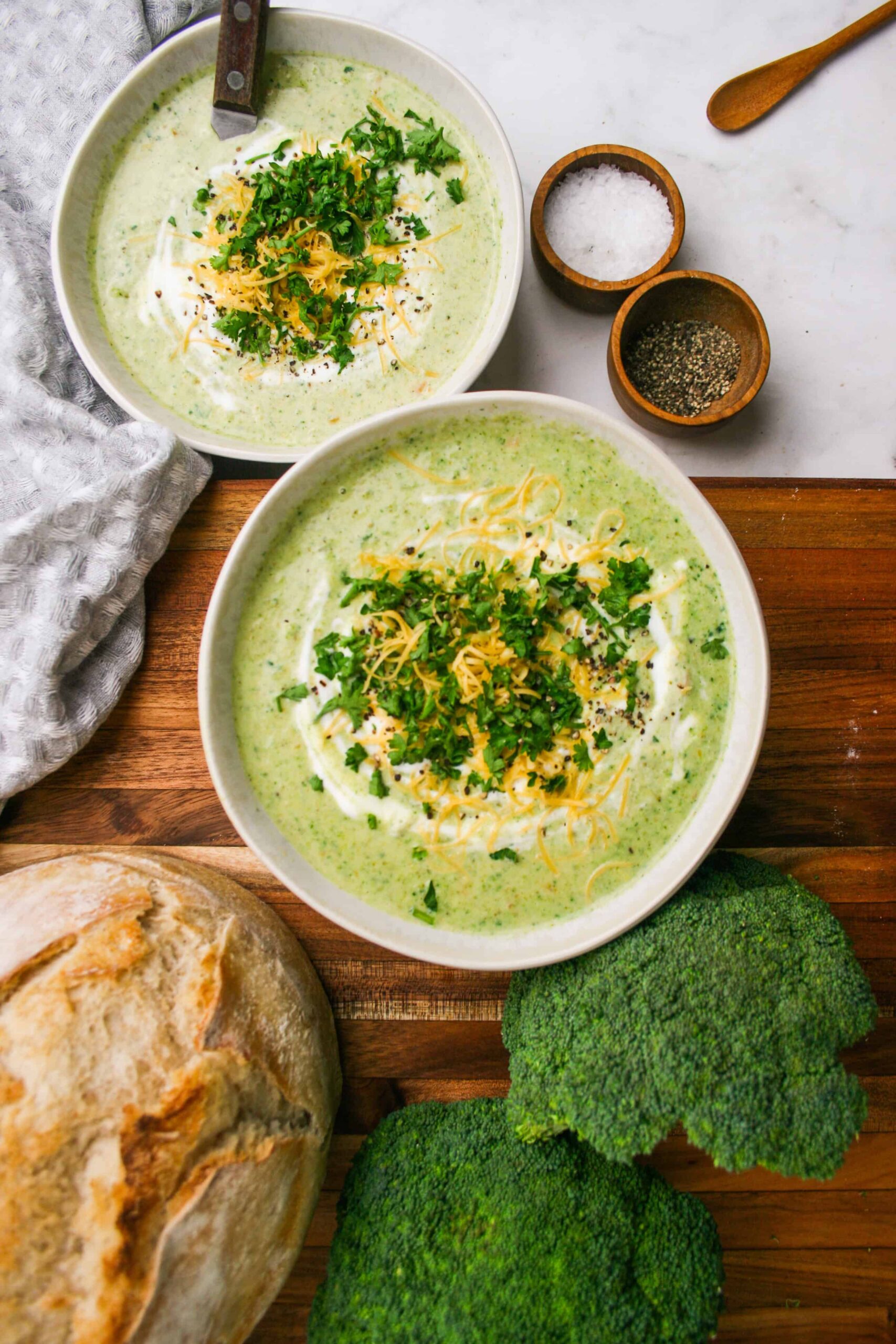 A rustic bowl of broccoli cheddar soup garnished with shredded cheddar and broccoli florets, photographed on a wooden table with warm natural light