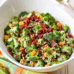 Overhead shot of a large white serving bowl filled with broccoli salad topped with crispy bacon bits, shredded cheddar cheese, sunflower seeds, and red onion, drizzled with creamy dressing, styled on a rustic wooden picnic table
