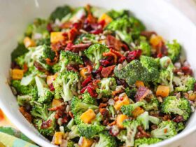 Overhead shot of a large white serving bowl filled with broccoli salad topped with crispy bacon bits, shredded cheddar cheese, sunflower seeds, and red onion, drizzled with creamy dressing, styled on a rustic wooden picnic table