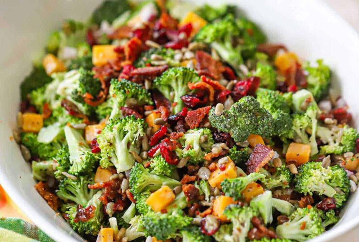 Overhead shot of a large white serving bowl filled with broccoli salad topped with crispy bacon bits, shredded cheddar cheese, sunflower seeds, and red onion, drizzled with creamy dressing, styled on a rustic wooden picnic table