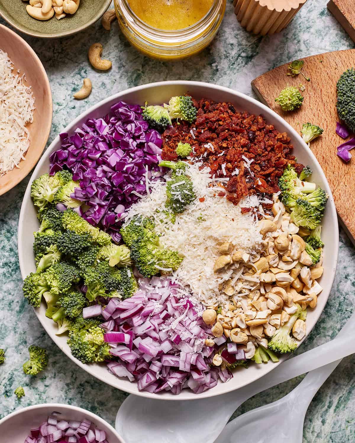 Flat lay of broccoli salad ingredients prepped and laid out — fresh broccoli florets, crispy bacon crumbles, shredded cheddar, sunflower seeds, dried cranberries, and diced red onion in small bowls on a light marble surface