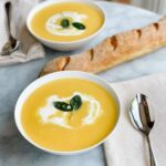 Silky butternut squash soup in a white bowl topped with a swirl of cream, crispy sage leaves, and cracked black pepper, photographed overhead on a rustic wooden table with autumn leaves