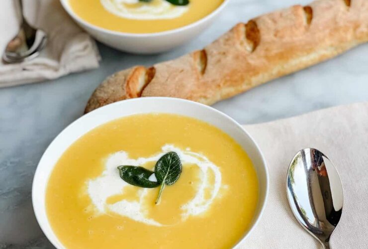Silky butternut squash soup in a white bowl topped with a swirl of cream, crispy sage leaves, and cracked black pepper, photographed overhead on a rustic wooden table with autumn leaves