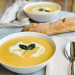 A stunning bowl of silky butternut squash soup garnished with a swirl of cream, crispy sage leaves, and a crack of black pepper, photographed overhead on a dark linen napkin with a rustic wooden spoon beside it
