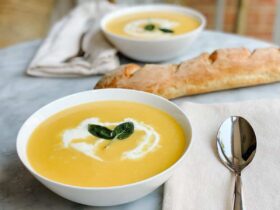 A stunning bowl of silky butternut squash soup garnished with a swirl of cream, crispy sage leaves, and a crack of black pepper, photographed overhead on a dark linen napkin with a rustic wooden spoon beside it