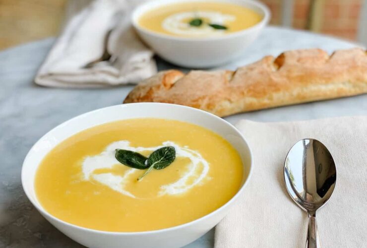 A stunning bowl of silky butternut squash soup garnished with a swirl of cream, crispy sage leaves, and a crack of black pepper, photographed overhead on a dark linen napkin with a rustic wooden spoon beside it