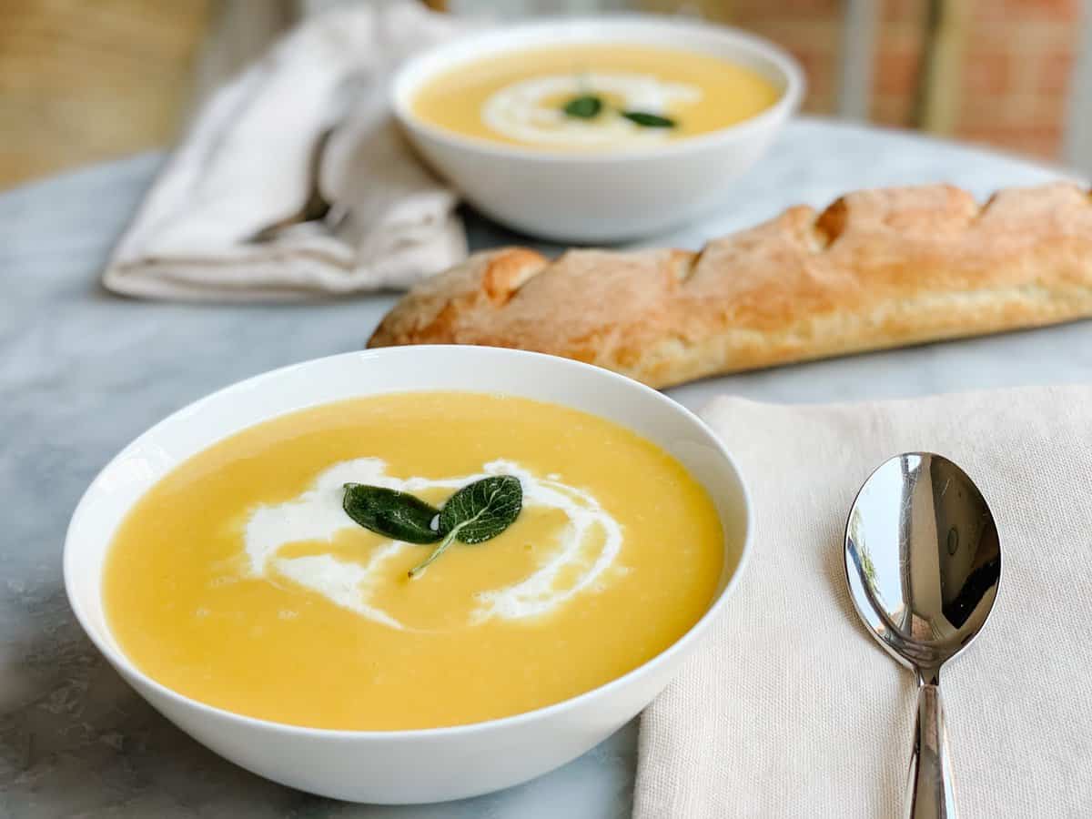 A stunning bowl of silky butternut squash soup garnished with a swirl of cream, crispy sage leaves, and a crack of black pepper, photographed overhead on a dark linen napkin with a rustic wooden spoon beside it