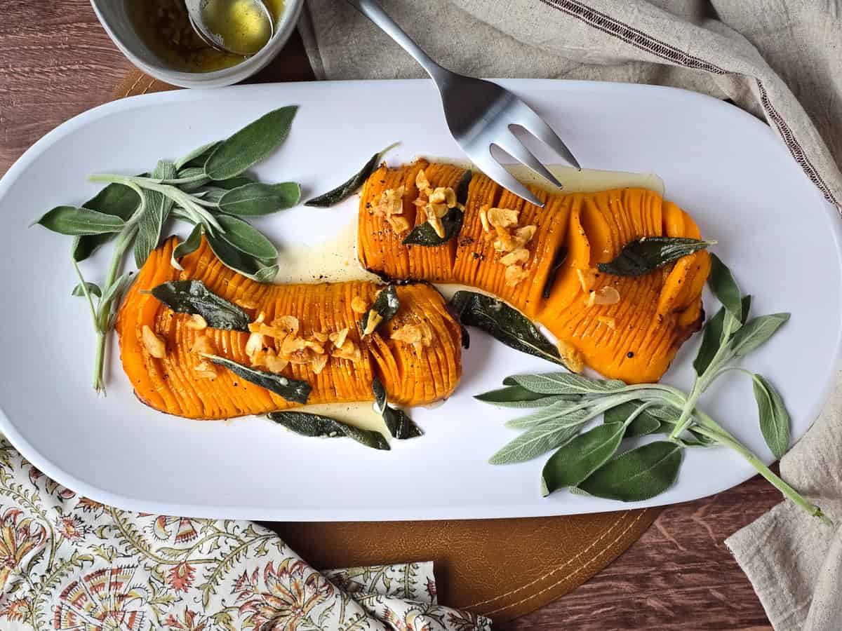 A flat-lay ingredients shot showing a halved butternut squash drizzled with olive oil on a sheet pan, alongside small bowls of sage, garlic, cream, and broth on a white marble surface